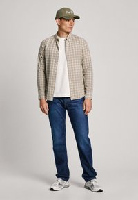 Camicia a maniche lunghe a quadri in beige e marrone, T-shirt bianca sotto, jeans blu e sneakers grigie. Cappellino oliva con dettagli testuali bianchi.