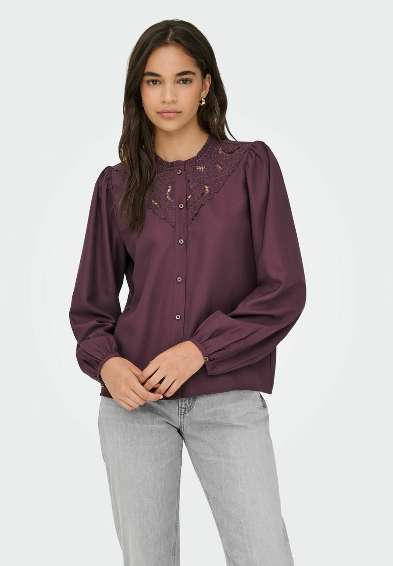 Vrouw met lang, donker haar, gekleed in een paarse blouse met knopen en kantdetails op de schouders, en lichtgrijze jeans, met de handen op de taille gekruist.