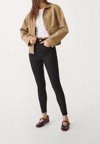 Chaqueta corta de cuero sintético marrón, top ajustado blanco, jeans skinny negros de tiro alto y bailarinas burdeos con una correa y acento dorado.