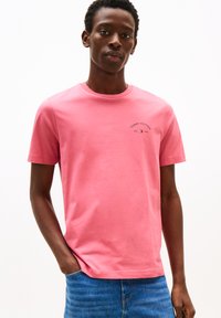 Homme portant un t-shirt rose Tommy Hilfiger et un jean bleu, se tenant avec une main dans la poche sur fond blanc.