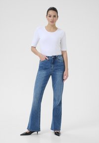 Mujer de pie con una mano en el bolsillo, vistiendo una blusa blanca de mangas cortas, jeans de tiro alto azules y tacones negros puntiagudos sobre fondo blanco.