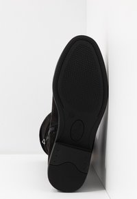 Semelle de botte noire avec motif antidérapant texturé, talon légèrement surélevé, et fermeture éclair latérale visible, positionnée contre des murs d'angle blancs.