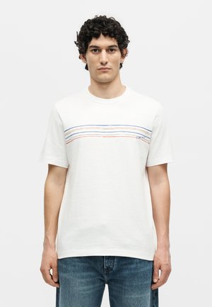 Νέος άνδρας με σγουρά μαλλιά που φοράει λευκή μπλούζα με οριζόντιες μπλε, πορτοκαλί και κόκκινες ρίγες και τζιν παντελόνι μπροστά σε απλό φόντο.