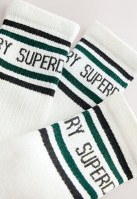 Hvide ribstrikkede sokker med sorte og grønne striber samt teksten "SUPERDRY" strikket nær manchetterne, vist overlappende på en lys baggrund.