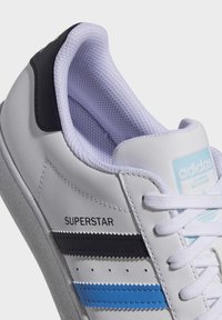 White leather sneaker with padded collar, blue and black stripes, rubber sole, and perforated detailing. Features "SUPERSTAR" label.