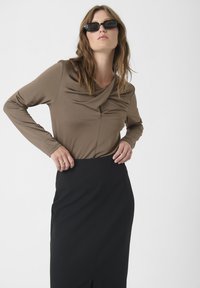 Haut long à manches brunes avec encolure drapée, associé à une jupe noire avec une fente sur le devant. Accentué par des lunettes de soleil noires.