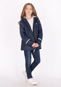 Jūros laivyno vandeniui atspari lietpalčio striukė su pilku interjeru ir atspindinčiomis detalėmis, derinama su mėlynomis džinsomis ir baltomis sportbačiais. Vaikas stovi su šypsena.