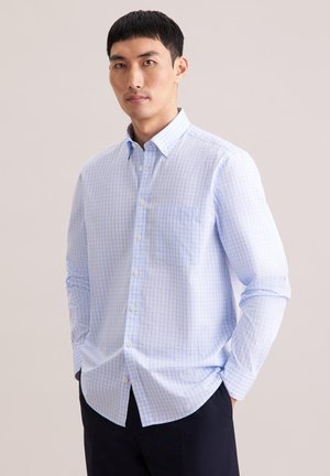 Uomo che indossa una camicia a maniche lunghe a quadri azzurri e pantaloni neri, in piedi davanti a uno sfondo semplice.