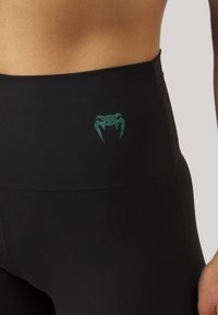 Leggings neri a vita alta in tessuto elasticizzato, con logo di ragno verde in vita e texture liscia.