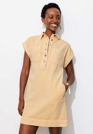 Femme souriante en robe beige à manches courtes boutonnée avec deux poches poitrine, boucles d'oreilles en forme de soleil et bracelet, debout devant un fond blanc.