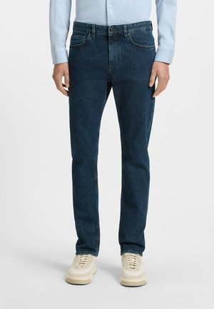 Dark blue denim jeans, slim fit, features front pockets and belt loops. Paired with light-colored sneakers, smooth texture.