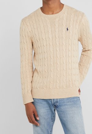 Maglione beige lavorato a maglia con motivo a trecce, scollo rotondo e polsini e bordo a coste. Piccolo logo sul petto. Abbinato a jeans azzurri chiari.