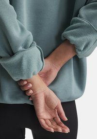 Lichtblauw sweatshirt met textuur en omslagmouw. De stof lijkt zacht, met een iets losse pasvorm. Handen zijn achter de rug gekruist.