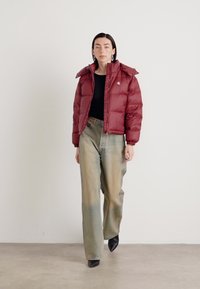 Giacca a vento bordeaux con colletto alto, texture lucida e polsini elasticizzati, abbinata a pantaloni larghi verdi sbiaditi e scarpe nere.