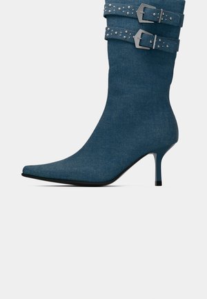 Blå denim kneehøye støvler med spiss tå, tynn hæl og dobbelt spennelåser med dekorative nagler. Glatt tekstur og strukturert design.