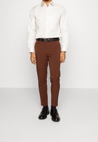 Camicia button-up beige chiaro abbinata a pantaloni marroni aderenti e scarpe eleganti nere. Include una cintura nera che rifinisce in vita.
