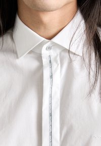 Primo piano di una persona che indossa una camicia bianca con colletto, con una striscia del logo "Michael Kors" lungo la pattina dei bottoni e lunghi capelli scuri.