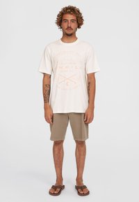 Cremfarbener T-Shirt mit orangefarbenem grafischen Druck und lockerer Passform, kombiniert mit beige Shorts. Modell trägt braune Sandalen und steht vor einem einfarbigen Hintergrund.