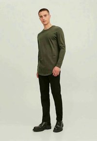 Jovem em pé, vestindo uma camisa de mangas compridas verde-oliva, calças pretas e sapatos pretos de atacadores, contra um fundo claro e simples.