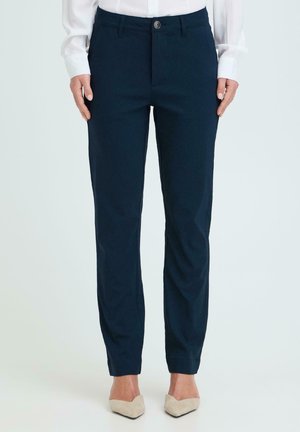 Femme portant un pantalon slim bleu marine et des chaussures pointues beige, debout les mains détendues le long du corps sur un fond clair.