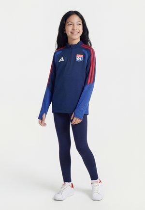 Pige med langt mørkt hår, der smiler, iført en blå og rød Olympique Lyonnais sportjakke, marineblå leggings og hvide sneakers på hvid baggrund.