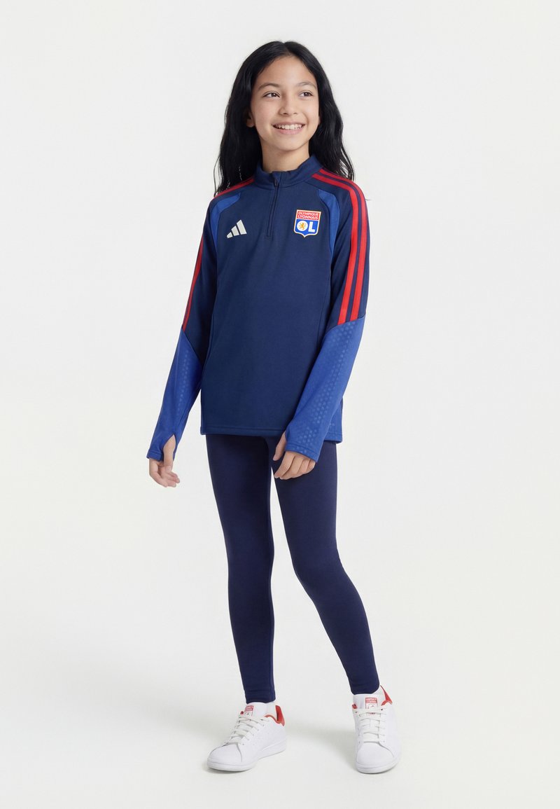 Fille aux longs cheveux foncés souriante, portant une veste de sport Olympique Lyonnais bleue et rouge, des leggings bleu marine et des baskets blanches sur fond blanc.