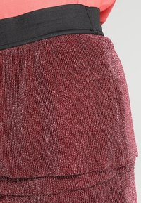 Jupe texturée rouge avec une finition scintillante, dotée d'une ceinture élastique noire. La surface présente un motif côtelé et un design en couches.