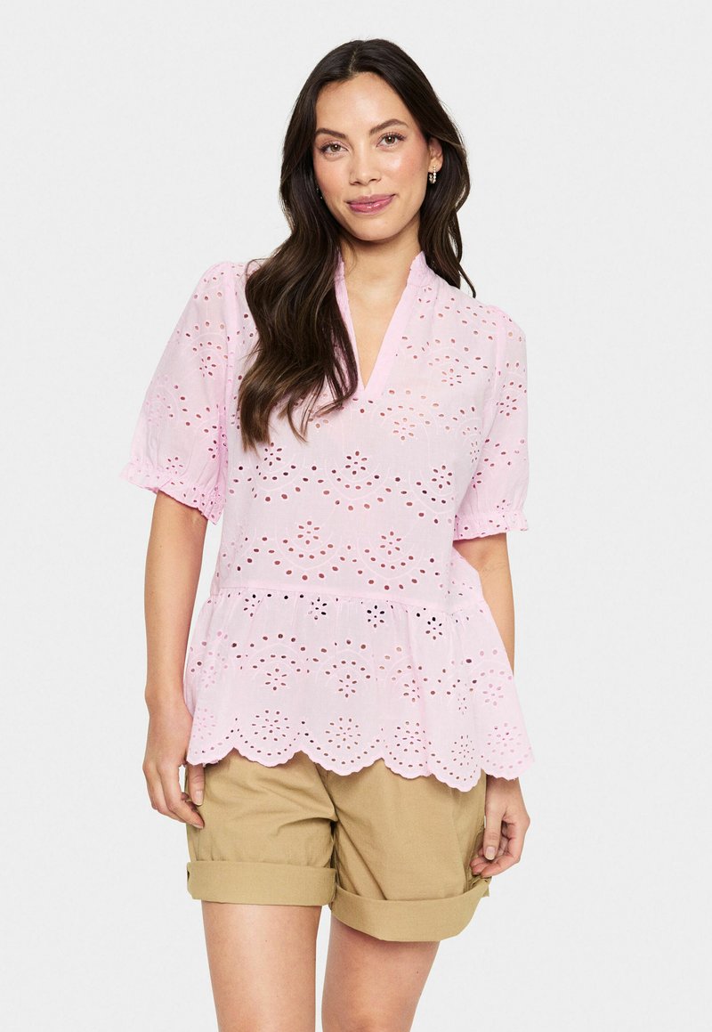 Rosa Augenblick-Bluse mit kurzen Ärmeln, gewelltem Saum und floralem Muster. Kombiniert mit braunen, hochgekrempelten Shorts.