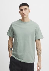 Világoszöld pamut póló rövid ujjakkal, kerek nyakkivágással és kényelmes fazonnal, egy tetoválásokkal rendelkező modellen bemutatva.