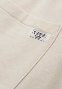 Close-up af beige stof med en hvid rektangulær label syet på, hvor der står "DIN ENESTE BEGRÆNSNING ER DIN HJERNE" og "OMBRE Siden 1985."