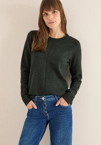 Maglione a coste verde scuro con scollatura rotonda, polsini a coste e dettaglio della cucitura centrale, abbinato a jeans blue a vita alta con chiusura a bottoni.