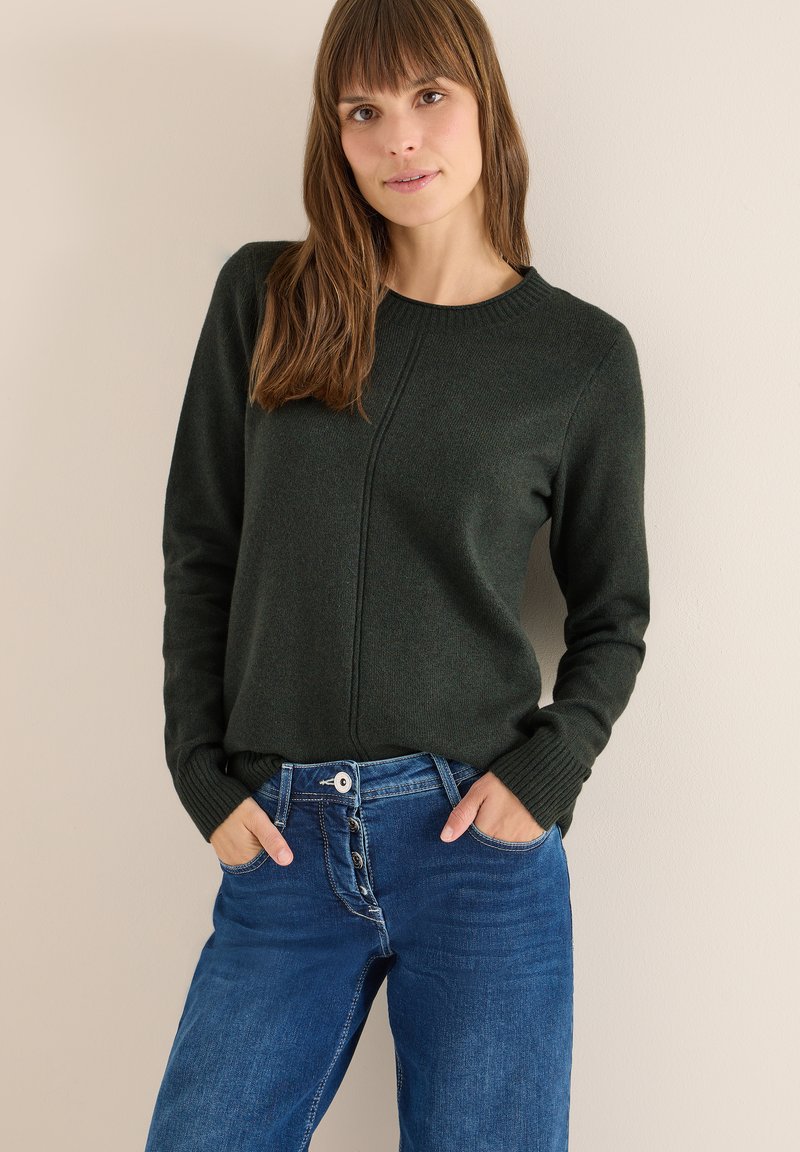 Maglione a coste verde scuro con scollatura rotonda, polsini a coste e dettaglio della cucitura centrale, abbinato a jeans blue a vita alta con chiusura a bottoni.