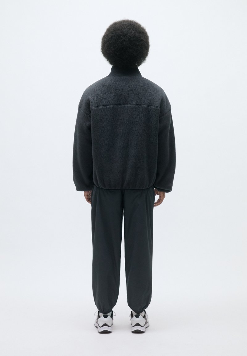 Svart fleecepullover med hög krage och sänkta axlar, kombinerad med mörkgrå smalbenade byxor. Sneakers med vit och svart sula.