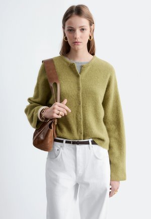 Jeune femme portant un cardigan boutonné vert olive, un pantalon blanc, des boucles d'oreilles anneaux dorés, et portant un sac à bandoulière en cuir marron.