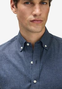 Knapp-up skjorta i mörkblått tyg med subtil textur och vita knappar på en spridd krage. Närbild av överkroppen.