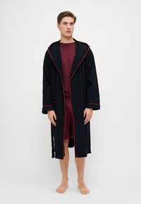 Mørkeblå badekåpe med teksturert stoff, rød piping langs kantene, hette-design og lange ermer, båret over et kastanjerødt antrekk.