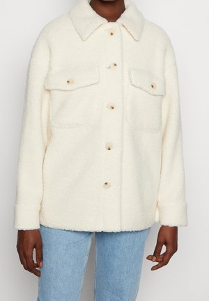Personne portant une veste crème, boutonnée, duveteuse avec deux poches poitrine et un jean bleu clair sur un fond uni.