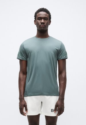 Rövid ujjú zöld sportpóló sima anyagból, lekerekített nyakkivágással és finom textúrával, fehér rövidnadrággal párosítva.