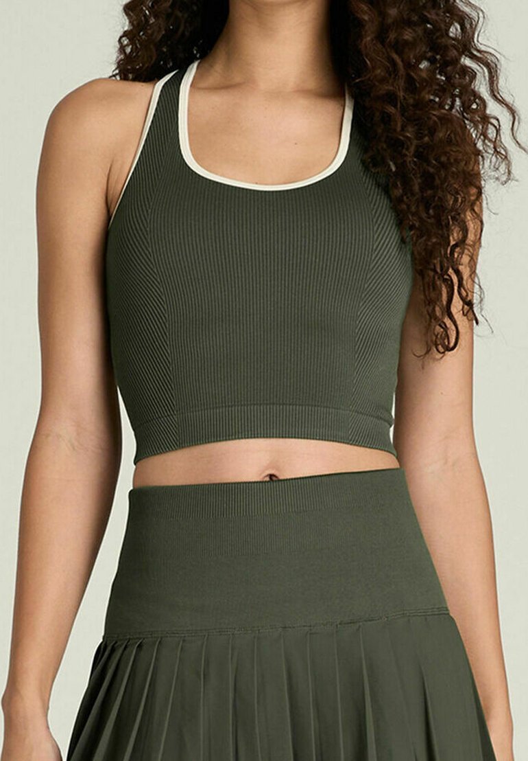 Frau trägt olivgrünes, geripptes Crop-Top mit weißen Kanten und passenden, hoch taillierten, plissierten Rock, dabei wird der Bauch gezeigt, und sie hat lockiges Haar.