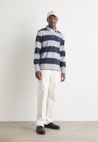 Tengeri kék és szürke csíkos galléros póló fehér gallérral. Fehér nadrággal, fekete cipővel és bézs színű, feliratos sapkával párosítva.