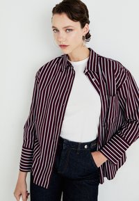 Črtasta srajca v maroon in beli barvi s kragljem nad navadno belo majico, v kombinaciji s temnimi denim skinny hlačami. Ima dolge rokave in žep.