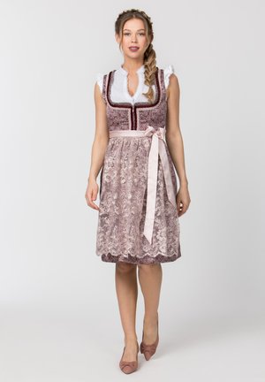 Frau trägt ein pinkfarbenes Spitzen-Dirndlkleid mit weißer Bluse und hellrosa Band, das in der Taille gebunden ist, und steht vor einem schlichten Hintergrund.