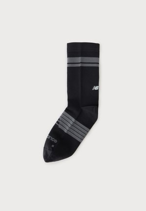 Czarna sportowa skarpetka z szarymi paskami blisko góry i stopy, z białym logo New Balance na łydce na białym tle.