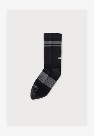 Chaussette de sport noire avec des rayures grises près du haut et du pied, avec un logo blanc New Balance sur la zone du mollet sur fond blanc.
