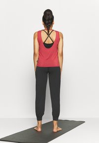 Rød tanktop med åben rygdesign over en sort sports-bh, parret med løse sorte joggingbukser, stående på en yogamåtte.