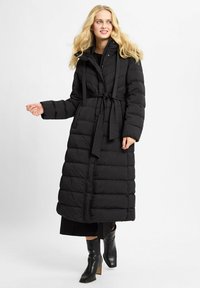 Blonde vrouw in een lange zwarte pufferjas met een riem, zwart jurkje en zwarte hoge laarzen, staande tegen een effen achtergrond.