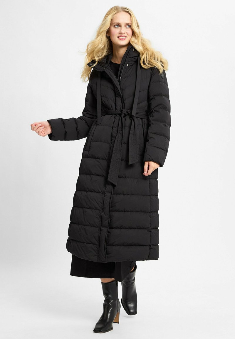 Blonde vrouw in een lange zwarte pufferjas met een riem, zwart jurkje en zwarte hoge laarzen, staande tegen een effen achtergrond.