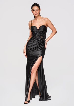 Kobieta w czarnej satynowej sukni wieczorowej z cienkimi ramiączkami, koronkowymi ozdobami i wysokim rozcięciem z przodu, nosząca czarne szpilki.