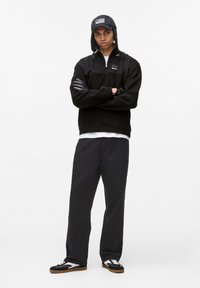 Pullover nero con collo a zip, sovrapposto a una camicia bianca, abbinato a pantaloni neri e sneaker in bianco e nero. Cappellino con dettaglio del logo.