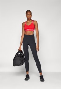 Sostén deportivo rojo con el logo "PRO" en blanco, leggings de talle alto negros, sosteniendo un bolso de gimnasio negro, usando zapatillas deportivas negras.